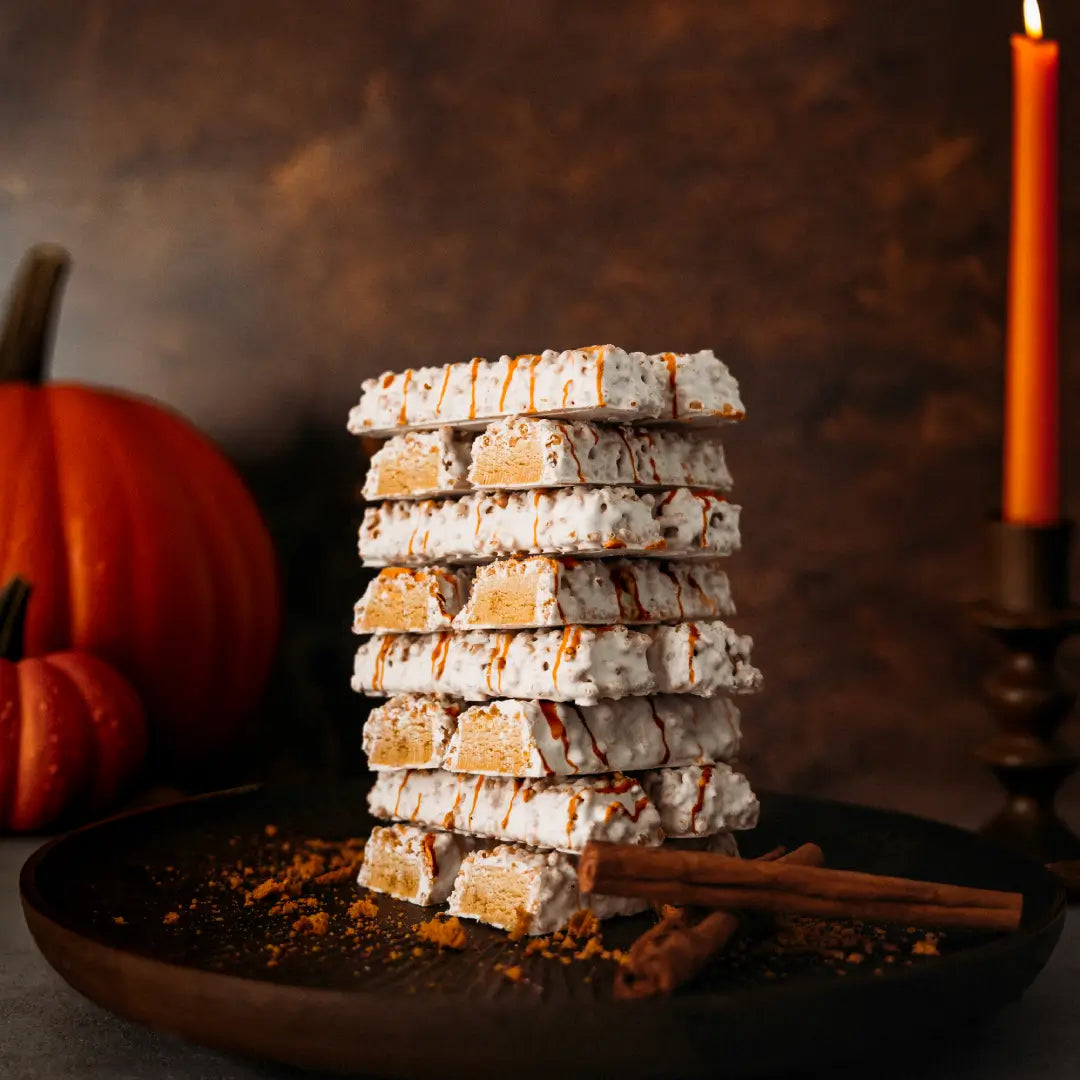 A stack of nine white chocolate-covered graham crackers drizzled with caramel, displayed on a dark plate with cinnamon sticks, a small pumpkin, and a lit orange candle in the background.