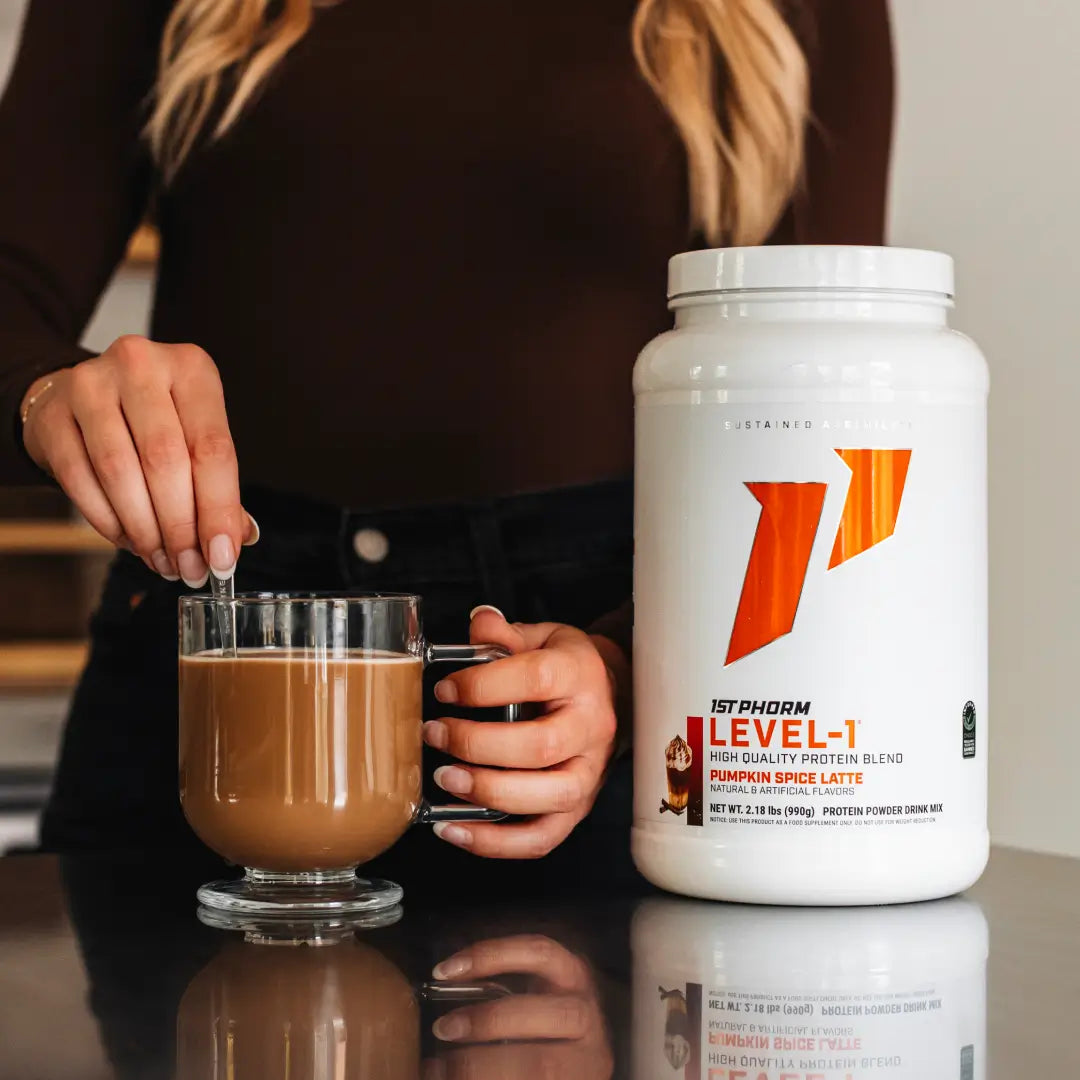 A person stirs a glass of coffee with a spoon beside a large white tub of Utopharm Level-1 High Quality Protein Blend in Pumpkin Spice Latte flavor, set on a reflective dark surface.
